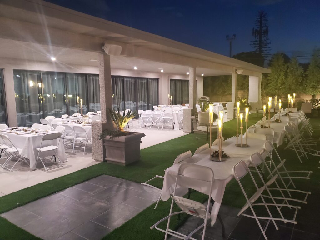 Outdoor event space set for dining with white tablecloths, chairs, and illuminated candle centerpieces, showcasing a welcoming evening atmosphere.