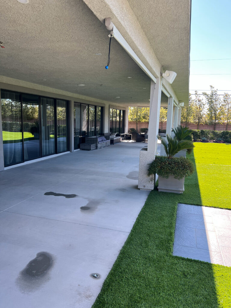 Covered patio with modern furniture, overlooking a well-maintained lawn. Visible water stains on the concrete floor add detail to the serene outdoor space.