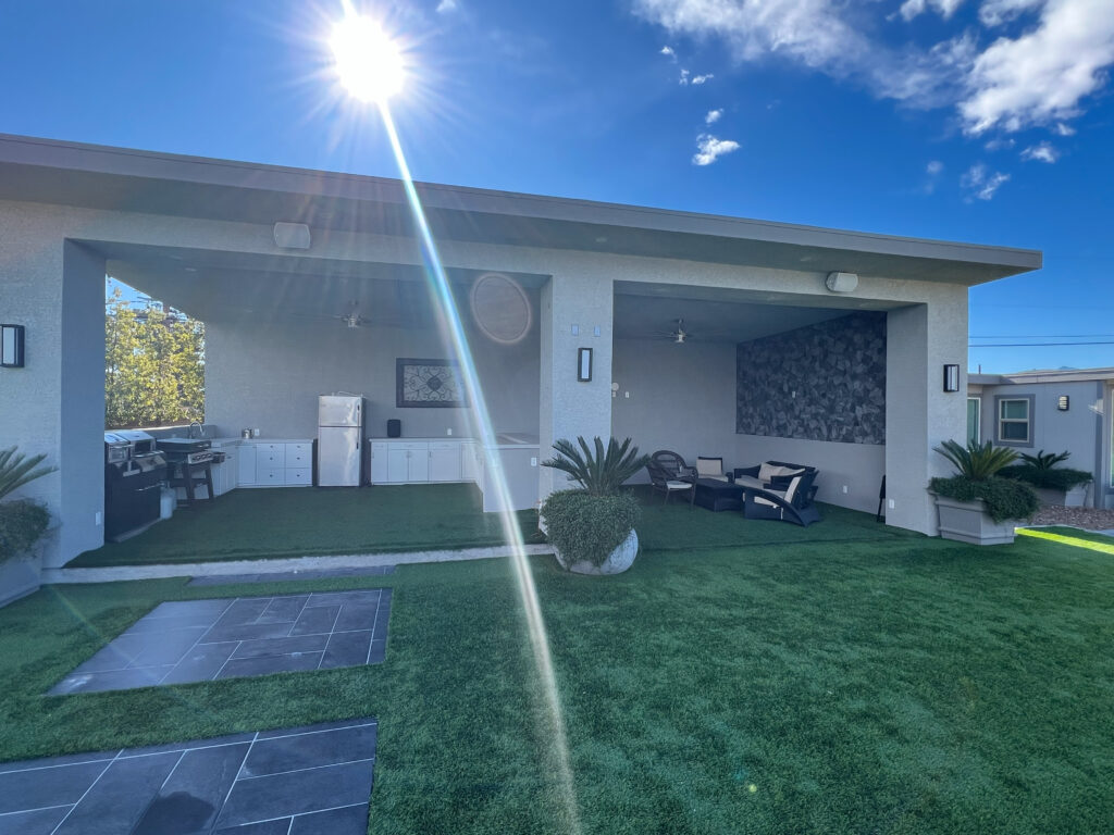 Outdoor entertainment area featuring a stylish kitchen with grill, adjacent seating space, and landscaped artificial grass under sunlight.