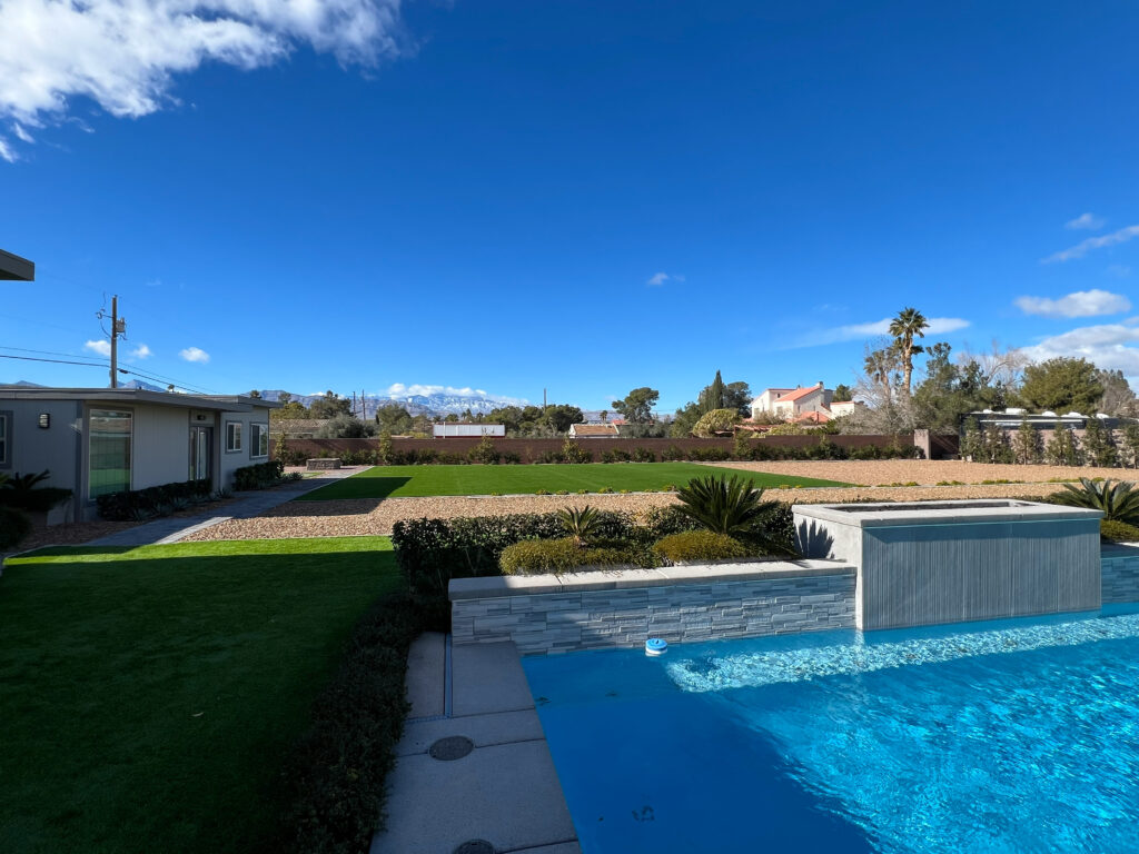 Modern backyard with a pool, landscaped grass, and desert plants, under a clear blue sky. Residences and mountains create a serene atmosphere.
