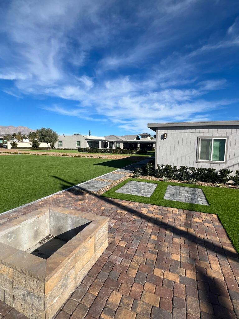 Patio scene featuring a stone fire pit surrounded by paver stones, with manicured lawn and modern buildings under a clear blue sky.