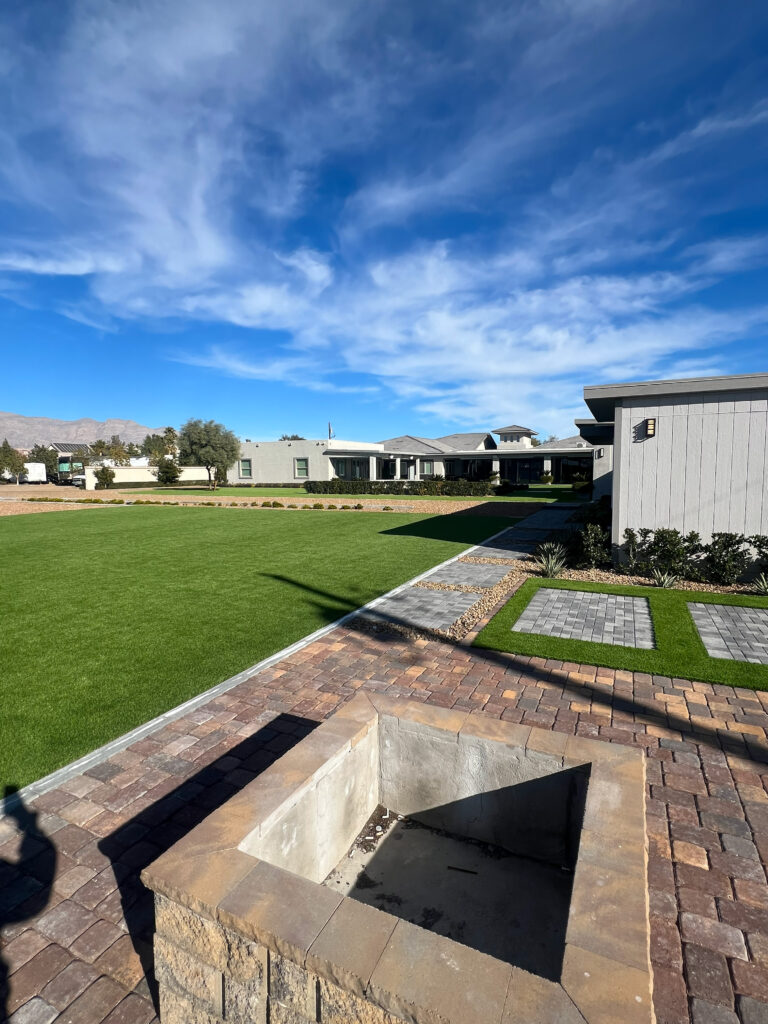Expansive green lawn under a clear blue sky, featuring a stone-bordered fire pit and modern landscaping in a residential area.