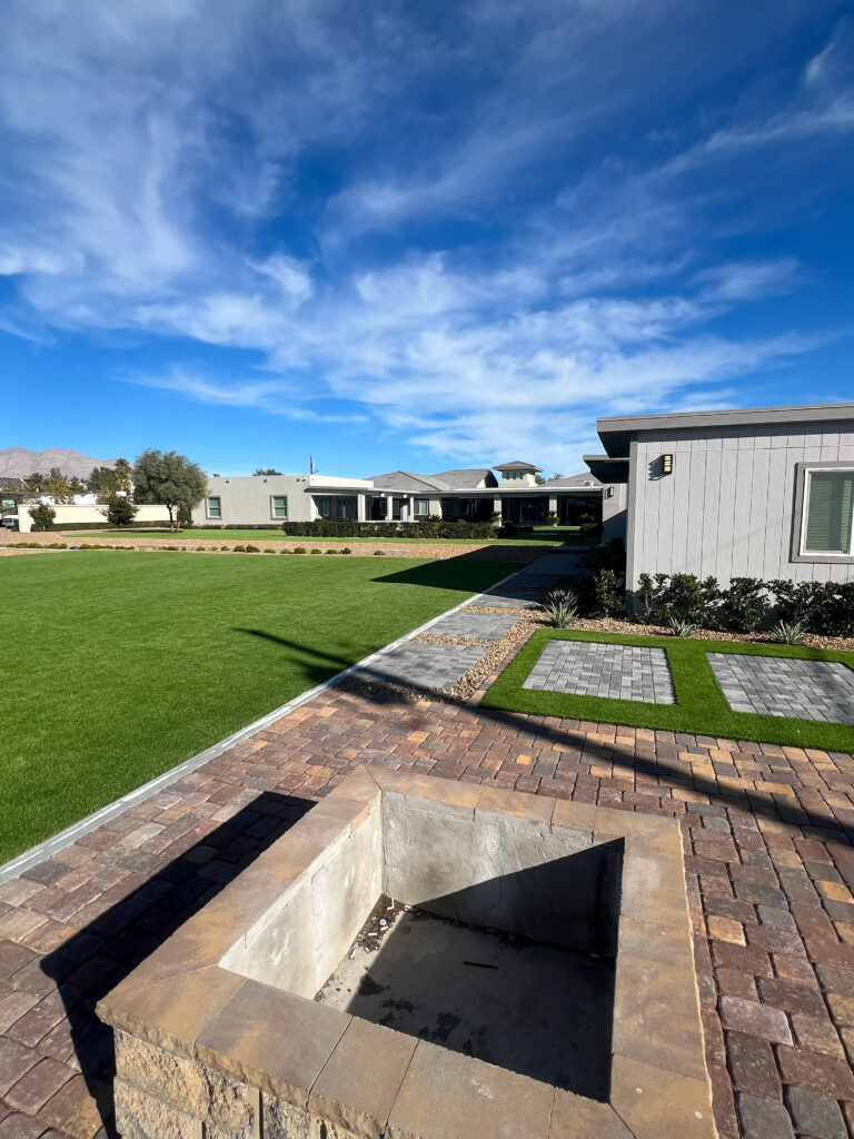 Concrete fire pit surrounded by paved stones and green grass, with modern houses in the background and a clear blue sky.