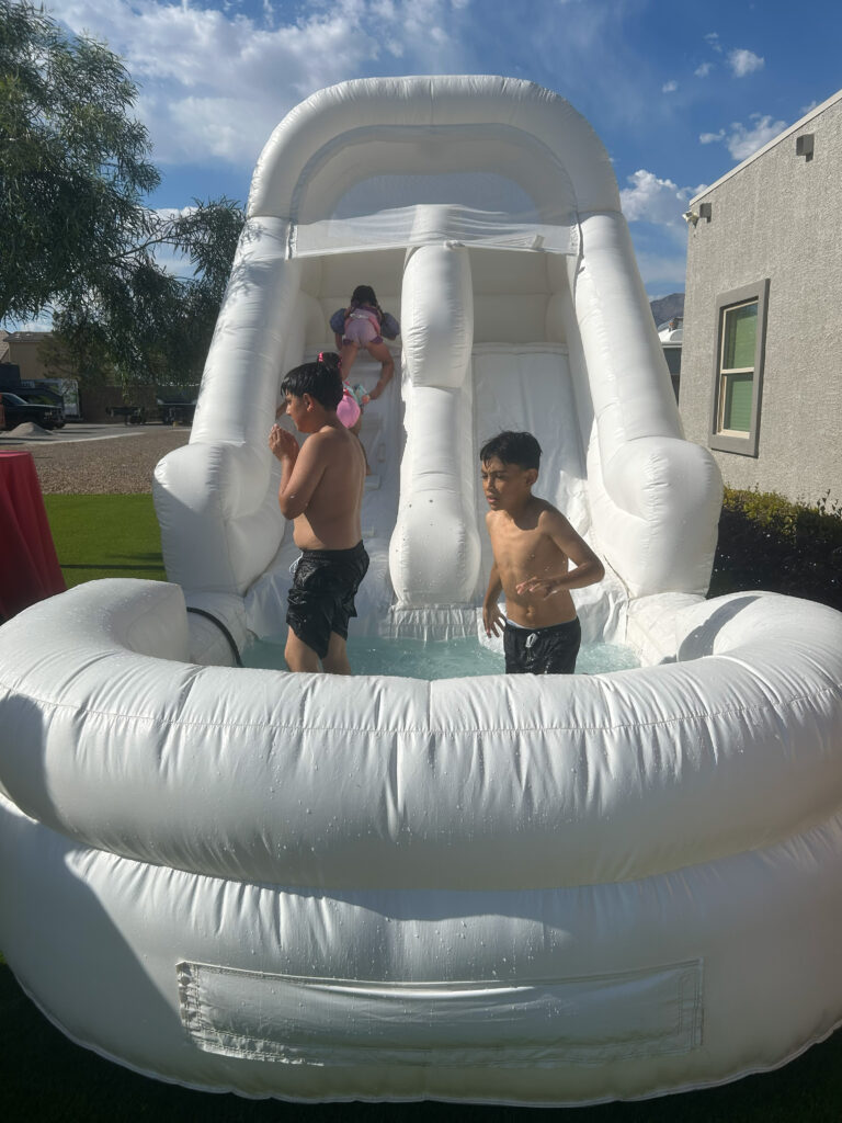 Kids enjoy a fun afternoon at a backyard water slide, splashing in the pool below while a girl climbs the slide in the background.