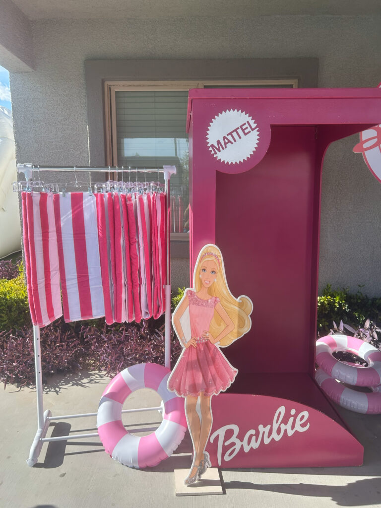 Barbie stands beside a pink booth displaying striped beach towels and lifebuoys, evoking a fun, summery atmosphere.