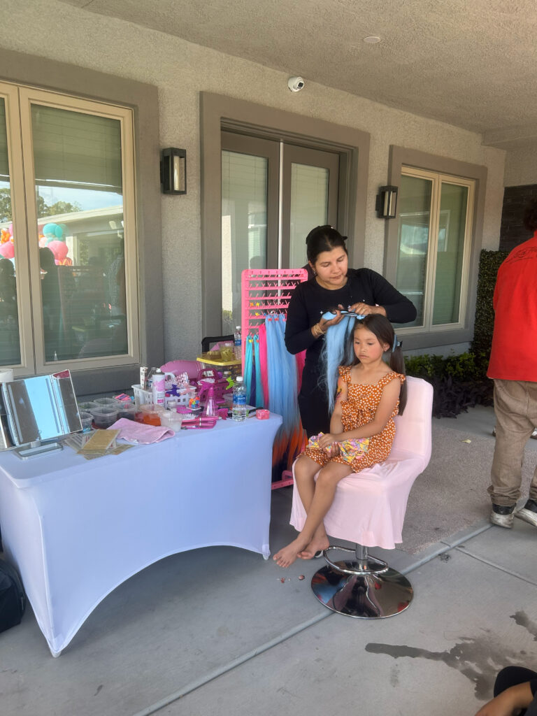 A hairstylist adds colorful extensions to a young girl's hair at an outdoor event, showcasing a playful atmosphere with decor in the background.