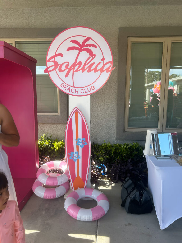 Sophia Beach Club sign features palm trees, a surfboard, and pink flotation rings, creating a festive beach-themed atmosphere for an event.