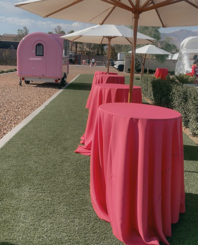 Pink tables with umbrellas line a lawn, leading to a pastel pink food truck in a lively outdoor event setting.