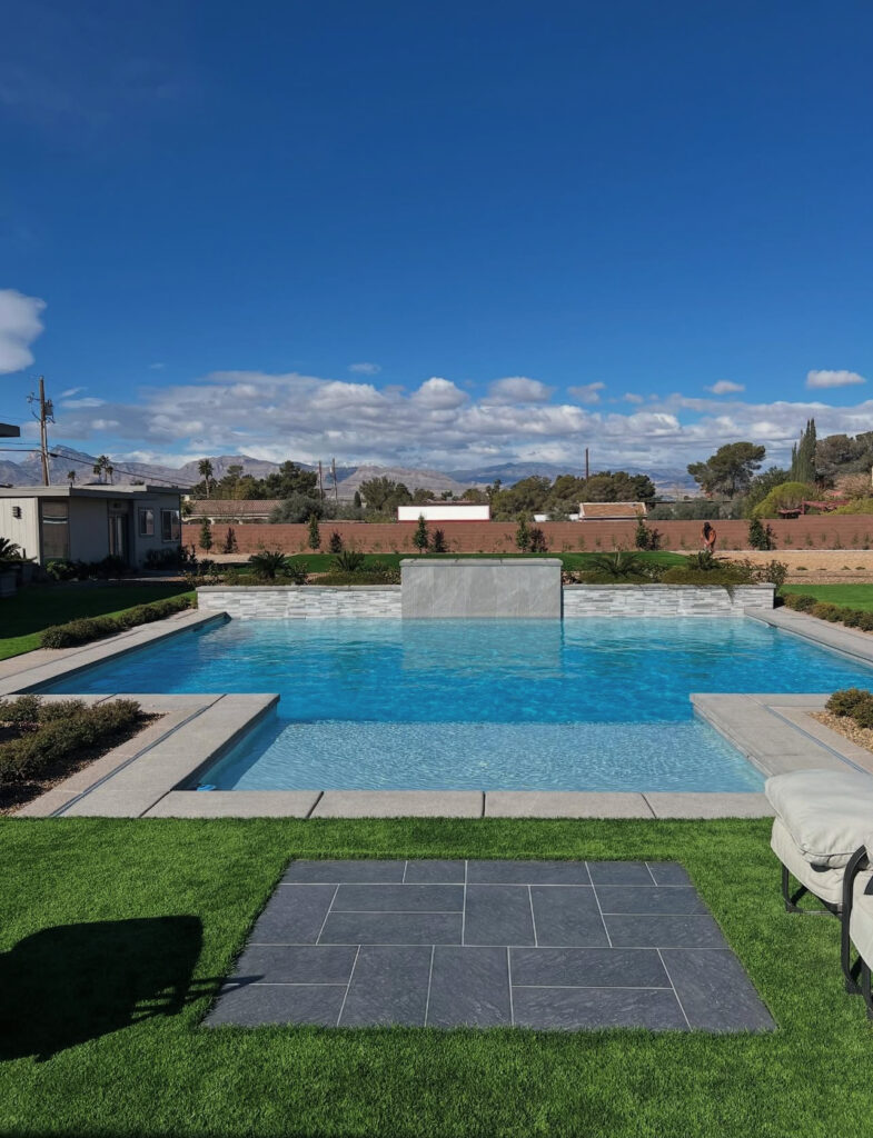 A tranquil backyard featuring a clear blue swimming pool with a stone water feature, surrounded by lush grass and plants under a bright sky.