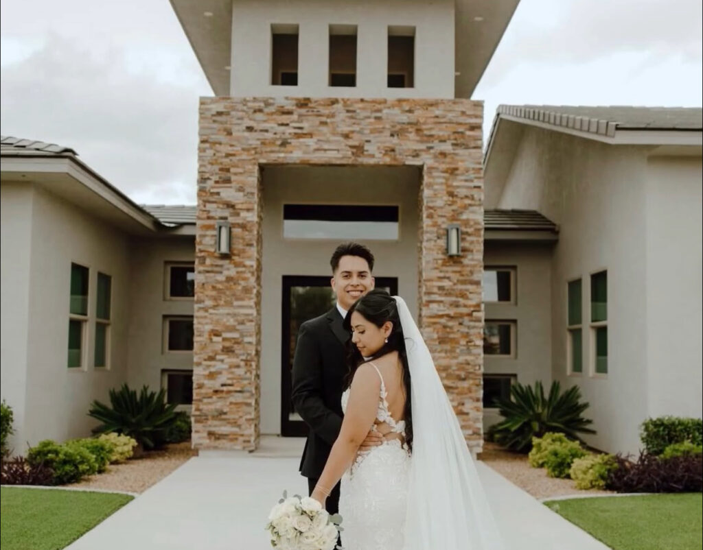 Newlyweds embrace in front of a modern home, showcasing the bride's elegant lace gown and bouquet, symbolizing a joyous celebration.
