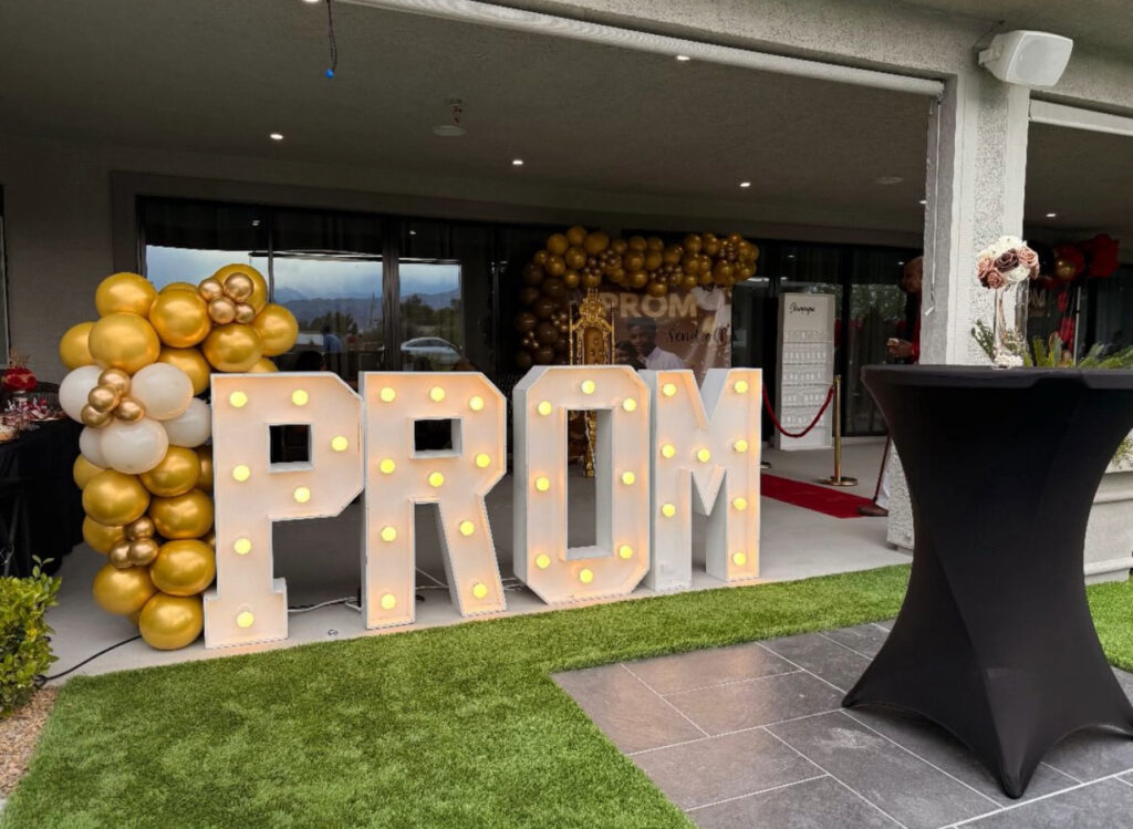 Illuminated "PROM" letters with gold and white balloons set the entrance for a prom event, featuring a red carpet and elegant decor.