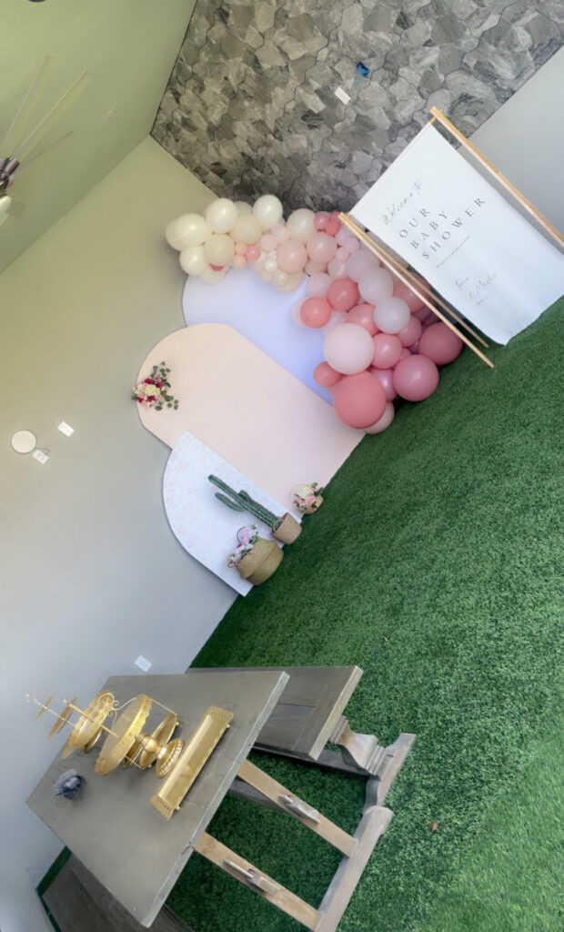 Decorative setup for a baby shower featuring a backdrop of pastel balloons, a welcome sign, and a stylish table on green turf.