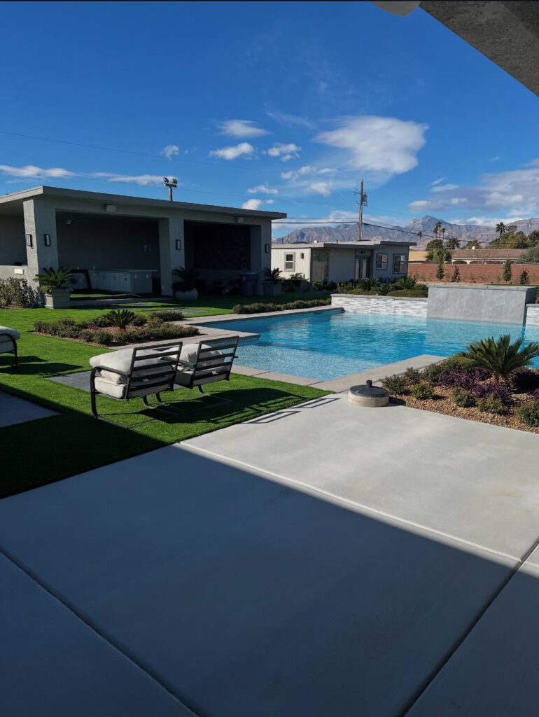 Modern outdoor area featuring a sparkling pool, lounge chairs, and landscaped greenery, with a mountain backdrop under a clear blue sky.