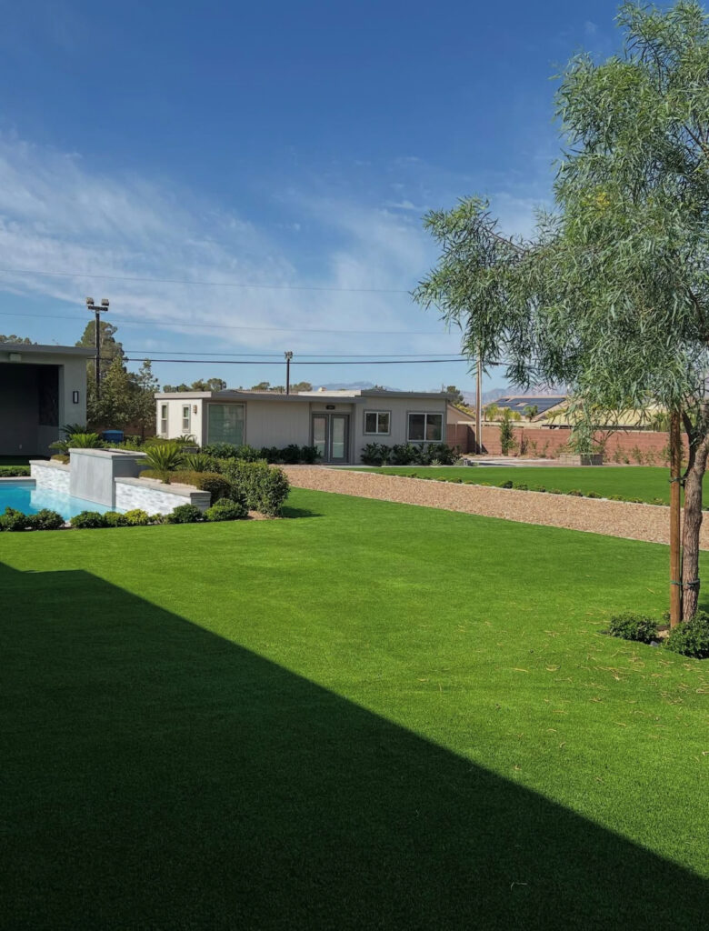 Lush green lawn with a swimming pool in the foreground, surrounded by modern homes and trees under a clear blue sky.