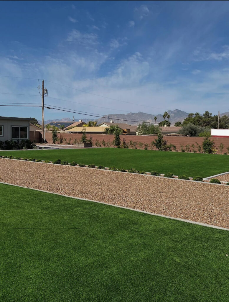 Lush green artificial lawn bordered by gravel pathways and shrubs, with distant mountains and a clear blue sky in the background.