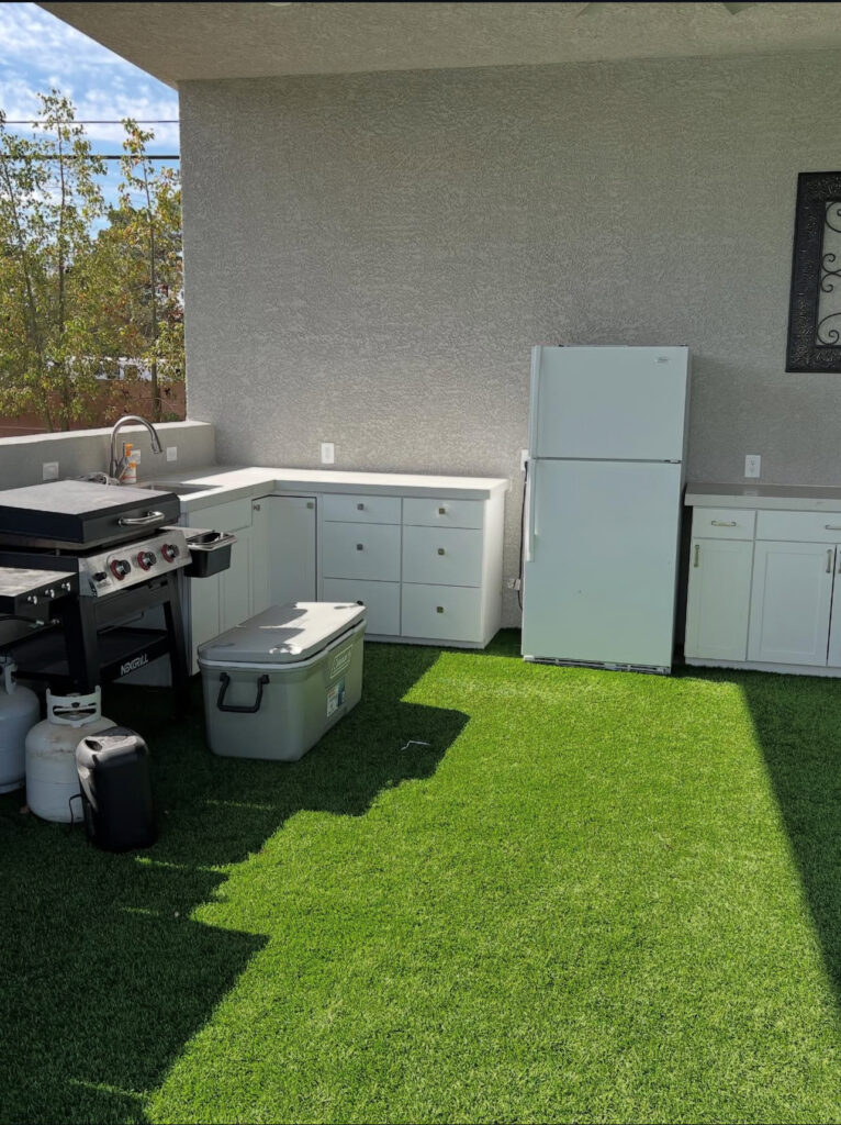 Outdoor kitchen featuring a grill, refrigerator, and storage cabinets on artificial grass. Ideal for cooking and entertaining in a backyard setting.