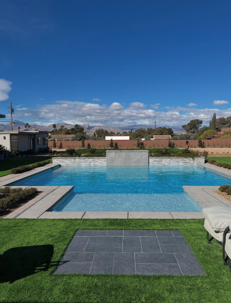 A modern swimming pool with a stone feature wall, surrounded by lush grass and landscaped gardens, under a clear blue sky.