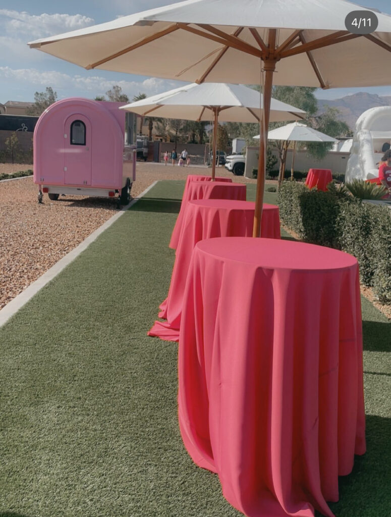 Pink tables under white umbrellas line a grassy path, with a pastel pink food trailer in the background, suggesting a festive outdoor event.