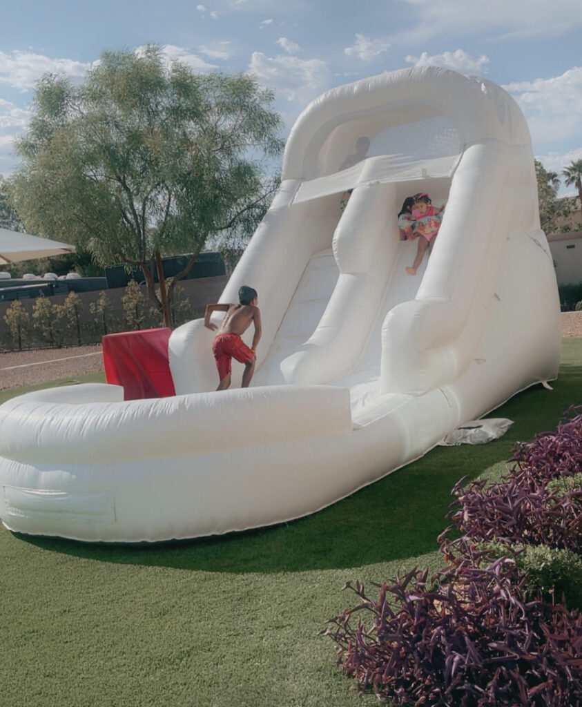 Children enjoy a large inflatable water slide, with one child climbing up as another prepares to slide down, set against a sunny backyard.