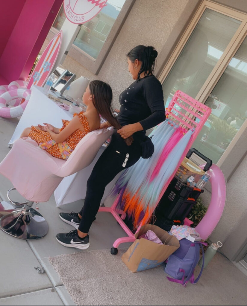A hairstylist braids the hair of a girl in a polka-dot dress, set in a vibrant beach club environment. Colorful hair extensions and beauty equipment are nearby.