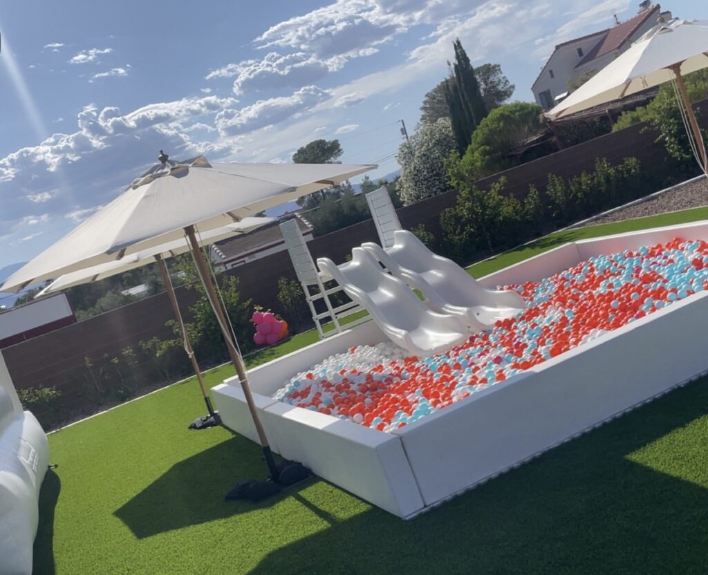 A vibrant backyard pool area featuring two white water slides and a colorful ball pit surrounded by umbrellas, ideal for summer fun.