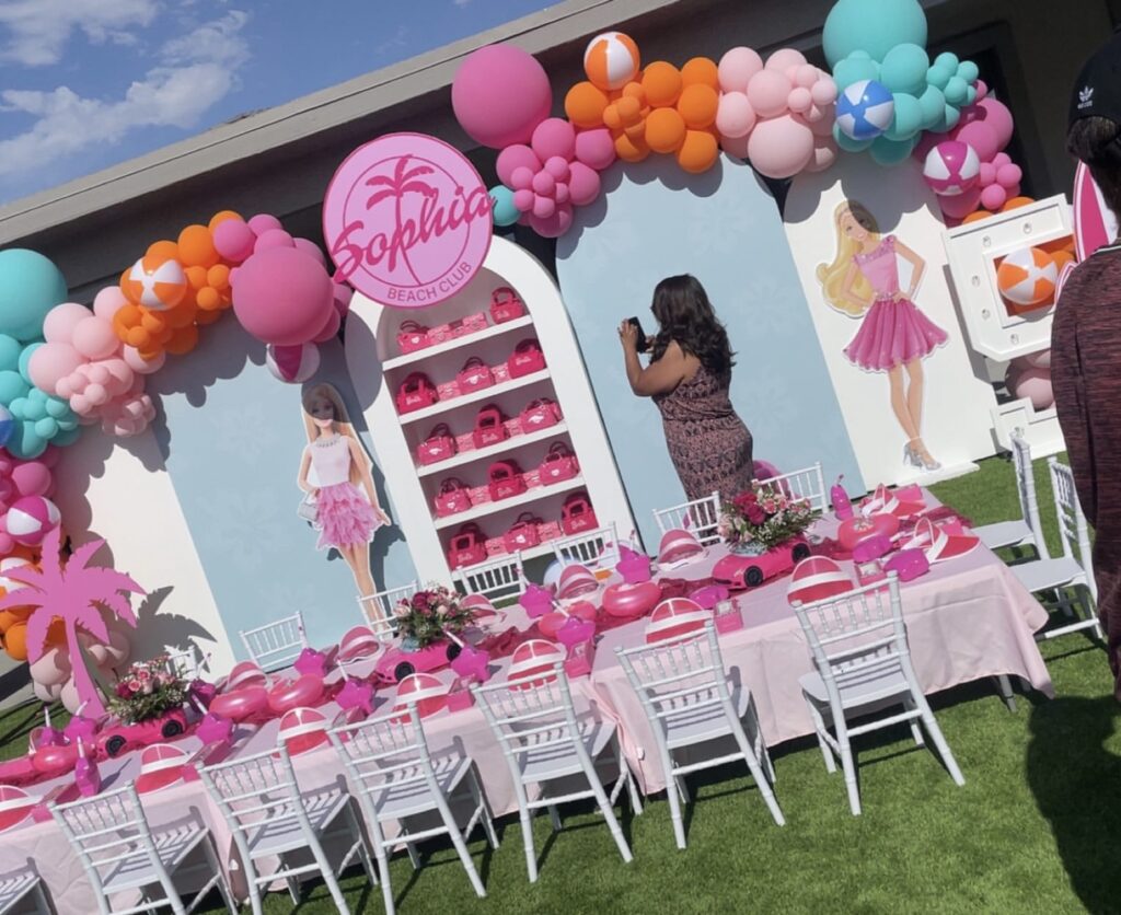 Colorful beach-themed party setup featuring a vibrant table, balloons, and Barbie decorations, celebrating "Sophia Beach Club." A woman captures photos.