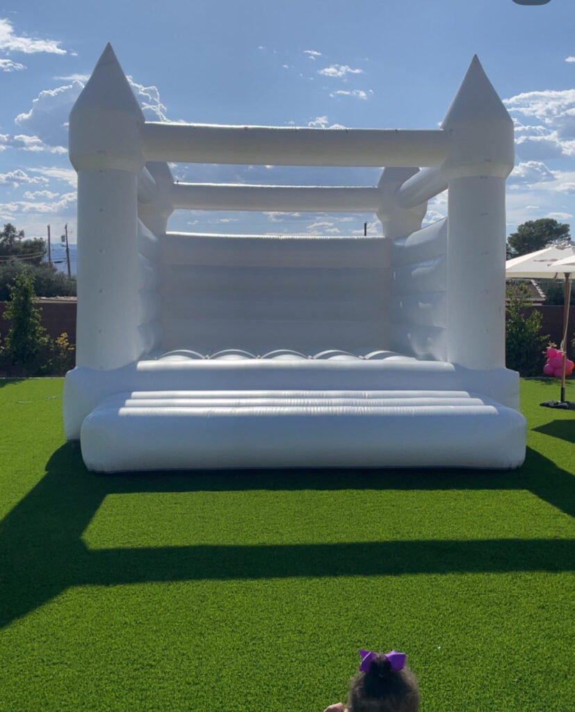 White inflatable bounce house with castle-like features, set on green artificial grass, under a bright blue sky. Ideal for children's events.