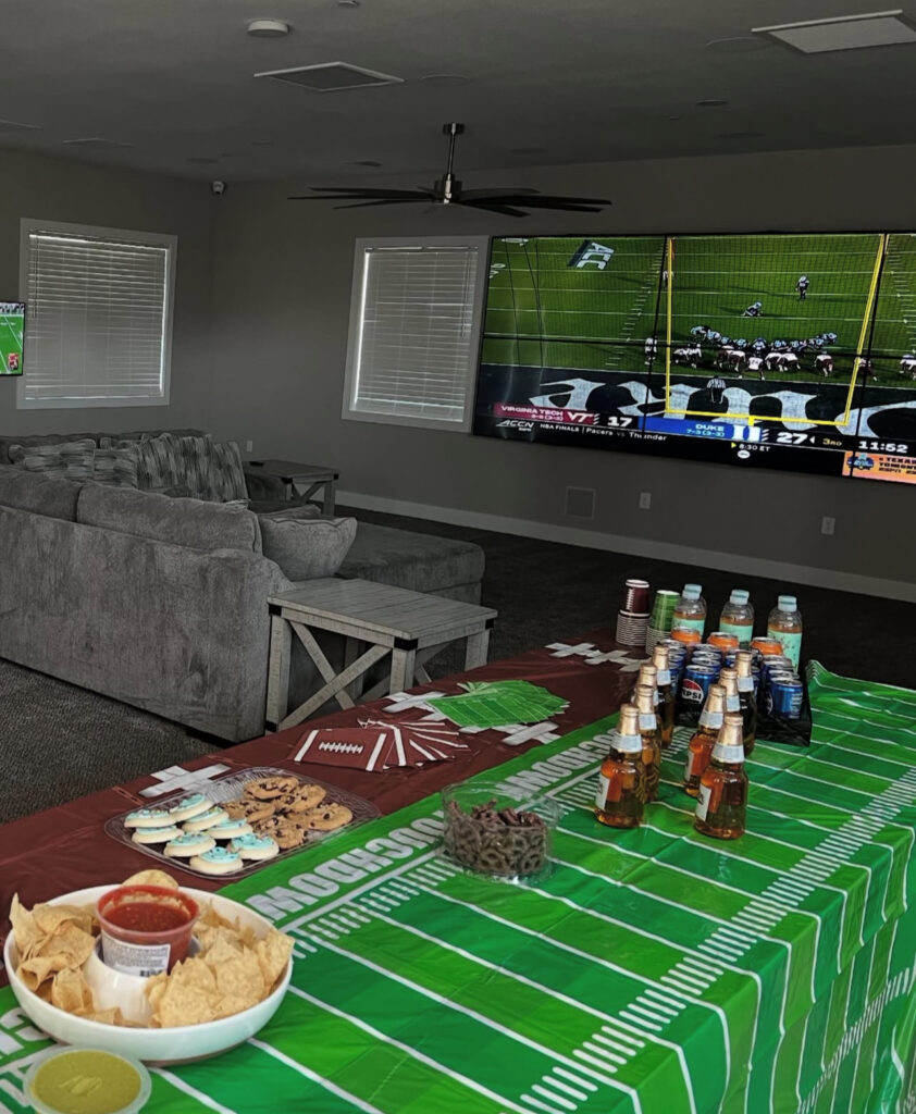 Living room set up for a sports gathering, featuring a large TV showing a football game, a snack table with chips, salsa, cookies, and drinks.