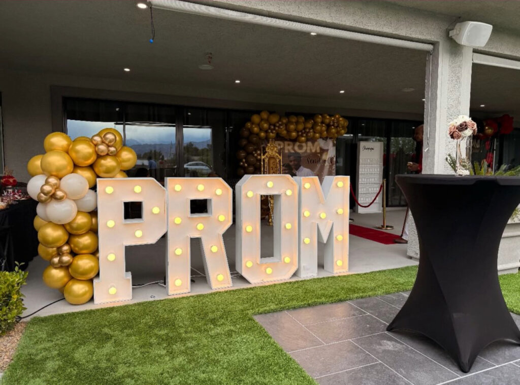 Illuminated marquee letters spell "PROM" at a festive outdoor venue, surrounded by gold and white balloons, setting a celebratory mood.