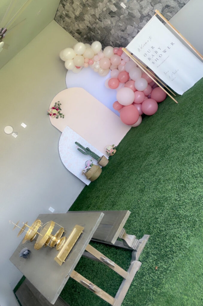 Decorative baby shower setup featuring pink and white balloons, a floral backdrop, and a wooden table with gold accents.