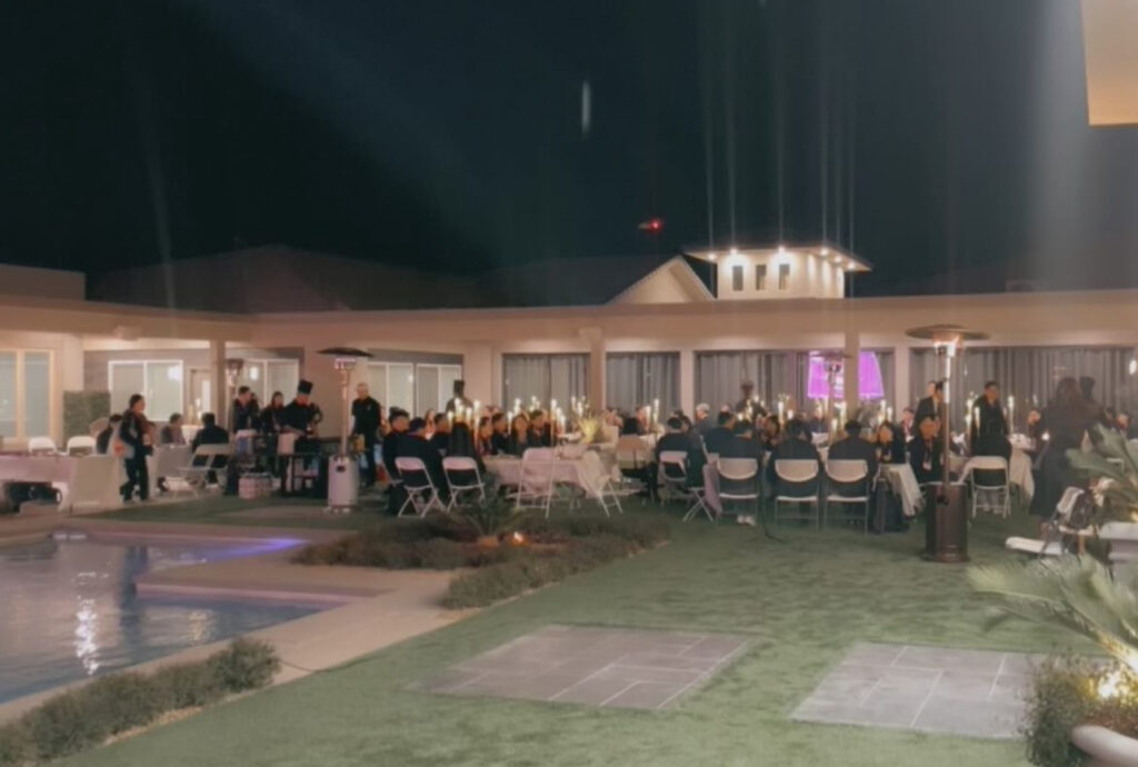 Outdoor evening event featuring a large gathering of guests seated at tables, illuminated by candles, with a pool and modern architecture in the background.
