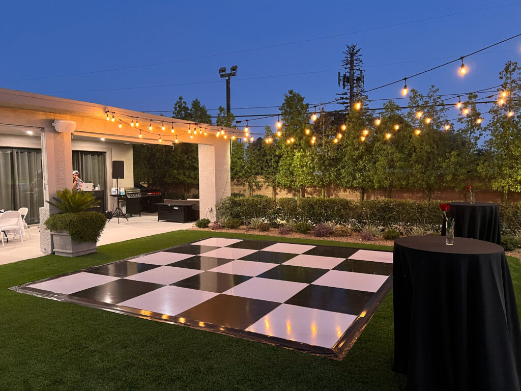 Outdoor event space at dusk featuring a large black-and-white checkered dance floor, string lights overhead, and two black cocktail tables with red roses.