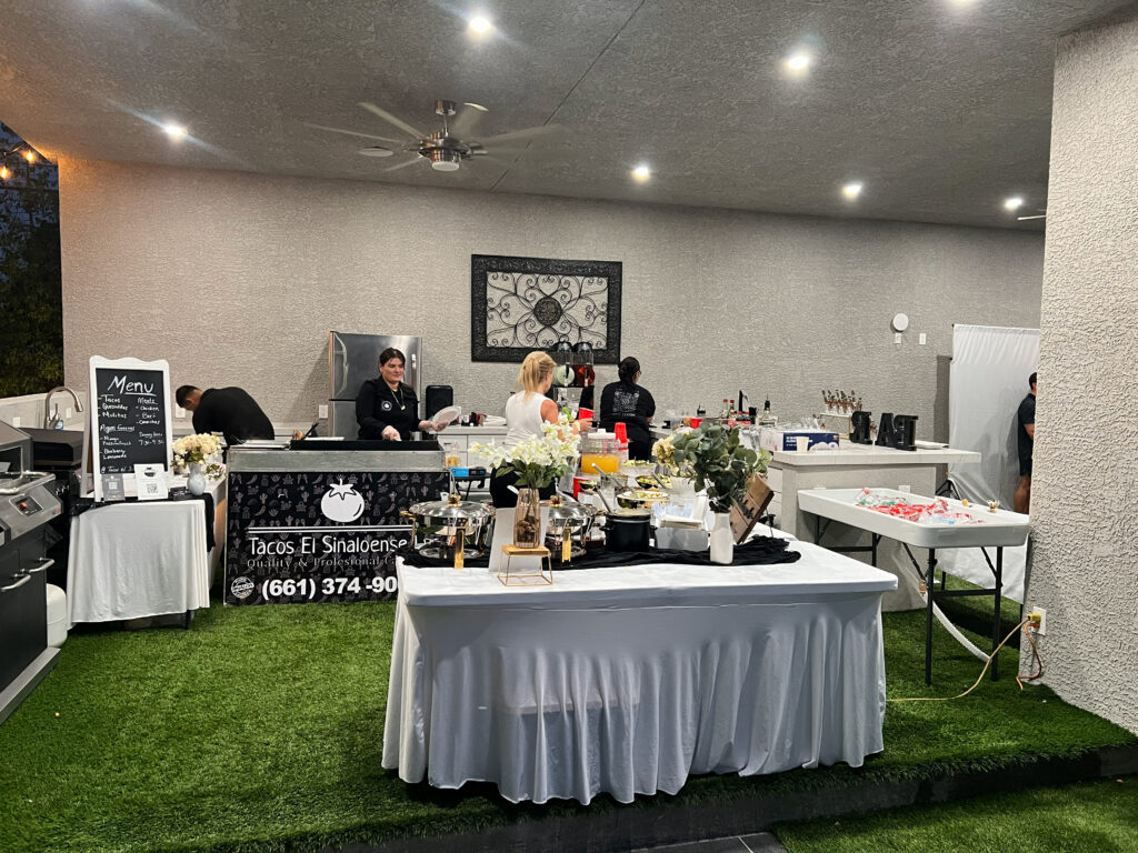 Catering setup at Tacos El Sinaloense features a menu board, food stations, and staff preparing dishes in a well-lit outdoor area.