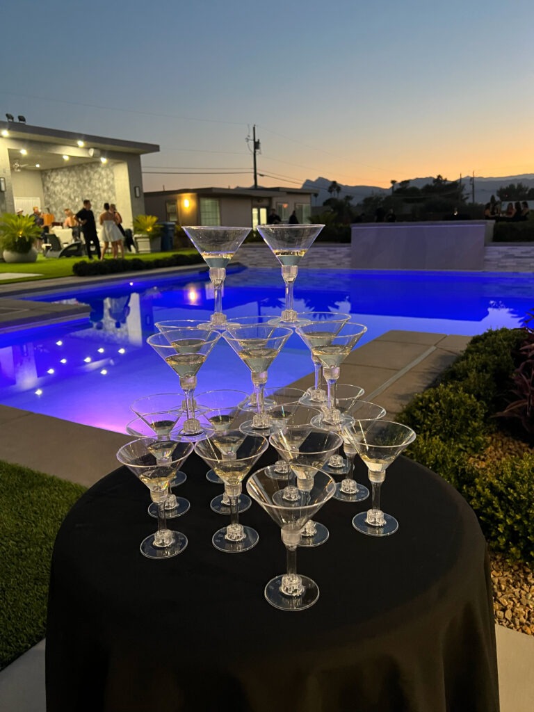 A pyramid of martini glasses sits on a black table by a glowing blue pool, with a modern house and guests enjoying the evening in the background.