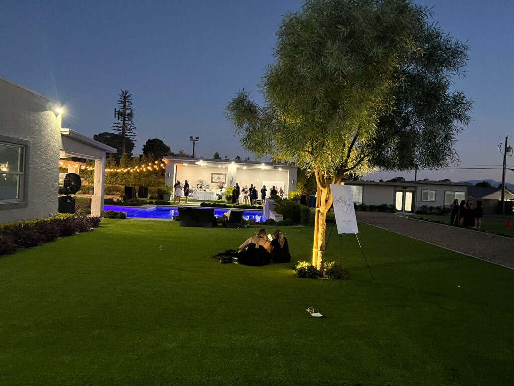A vibrant outdoor venue at dusk, featuring a well-lit lawn, a glowing pool, and guests mingling near a decorated area for events.