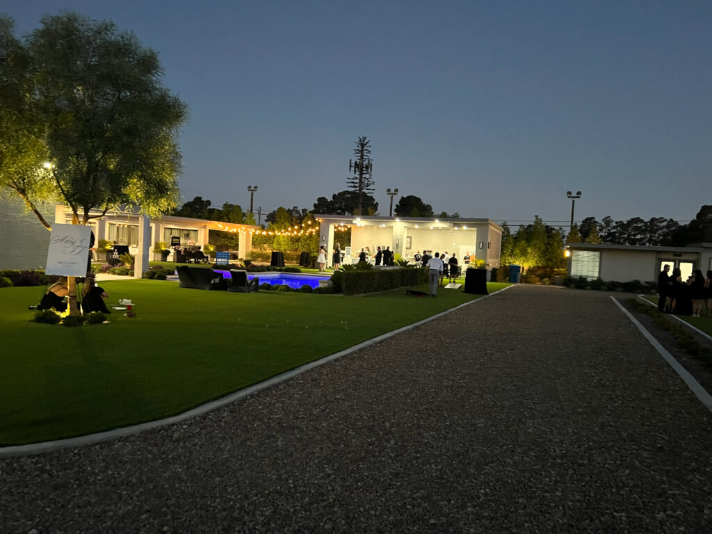Elegant outdoor event featuring a lit patio with guests mingling by a pool. The setting is adorned with greenery, creating a festive atmosphere.