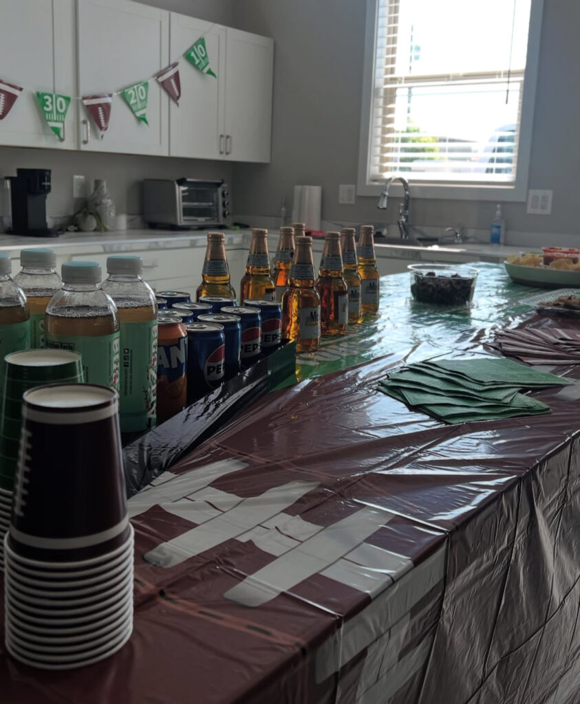 A festive kitchen counter displays a variety of beverages, including bottled drinks, sodas, and snacks, with football-themed decorations overhead, ideal for a game day gathering.