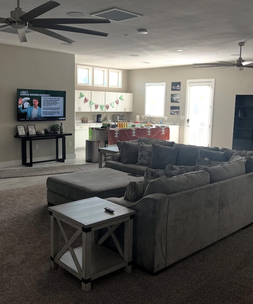Cozy living room with a large gray sectional sofa and a wooden side table. A TV displays career accomplishments, while a festive kitchen setup is visible.