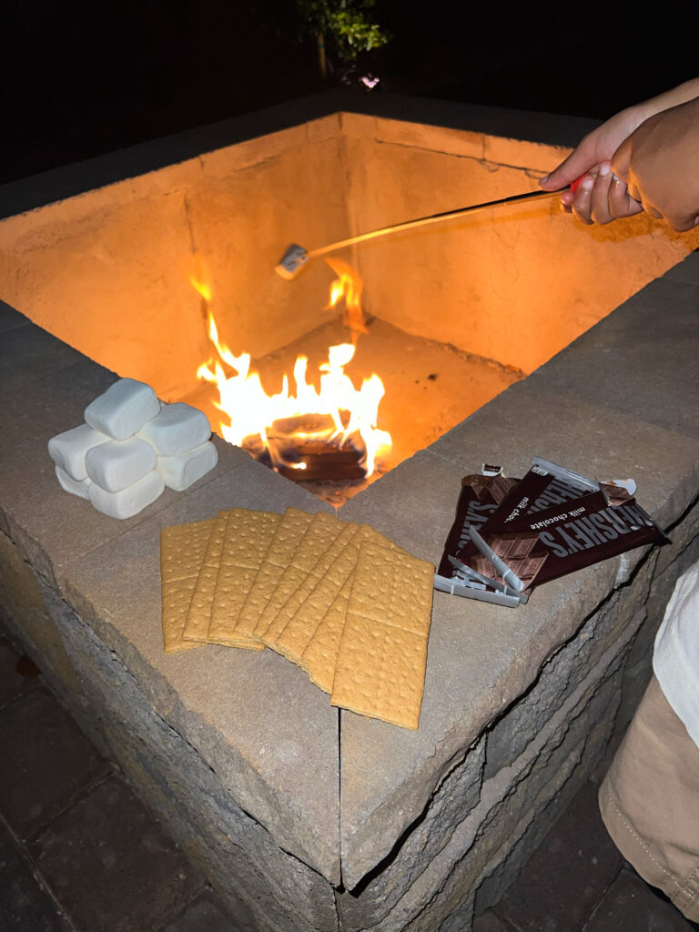 A person roasts a marshmallow over a fire pit, with graham crackers and chocolate nearby, preparing for s'mores in the evening.